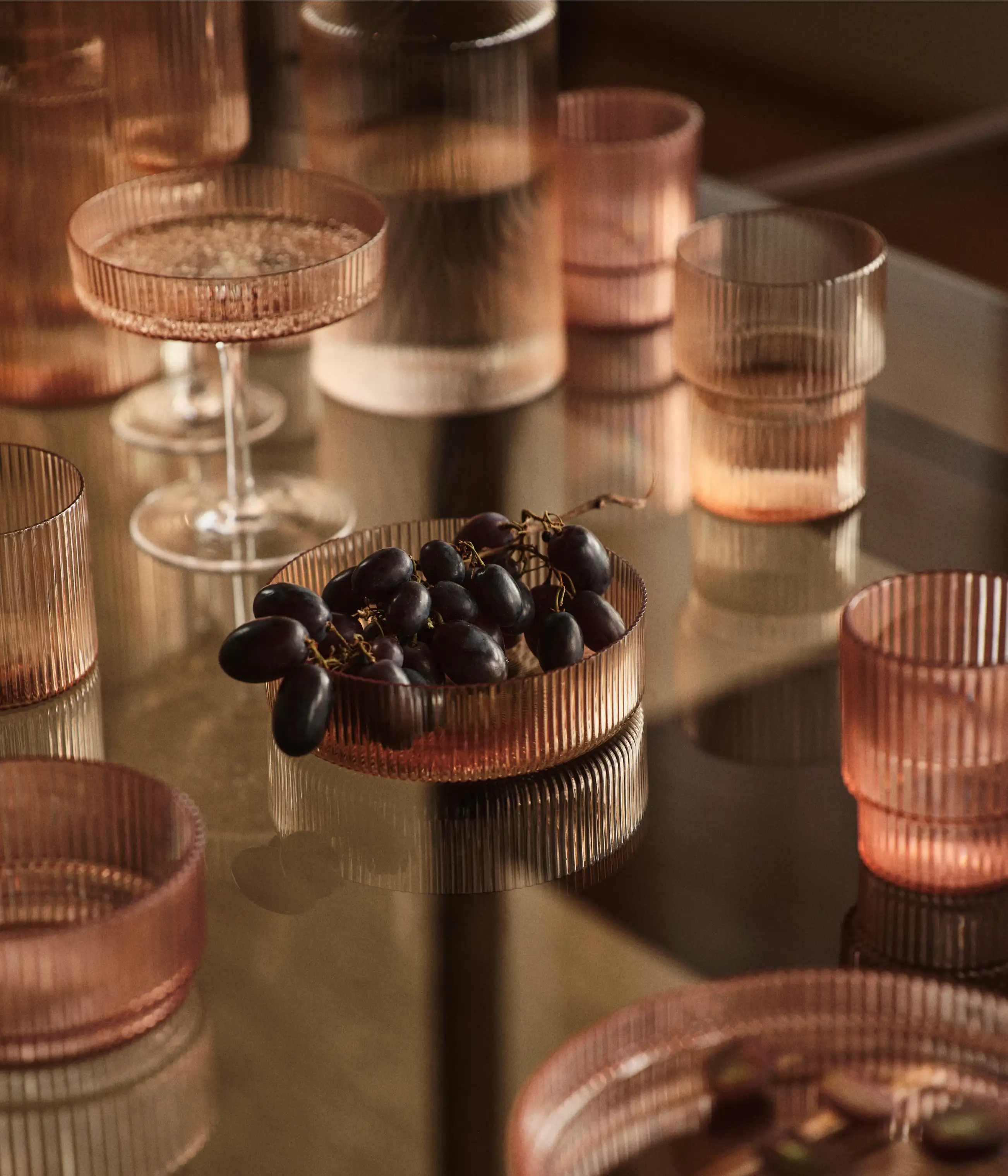 Riflet glas på et spejlbord, skål med mørke druer, vandglas og et coupeglas
