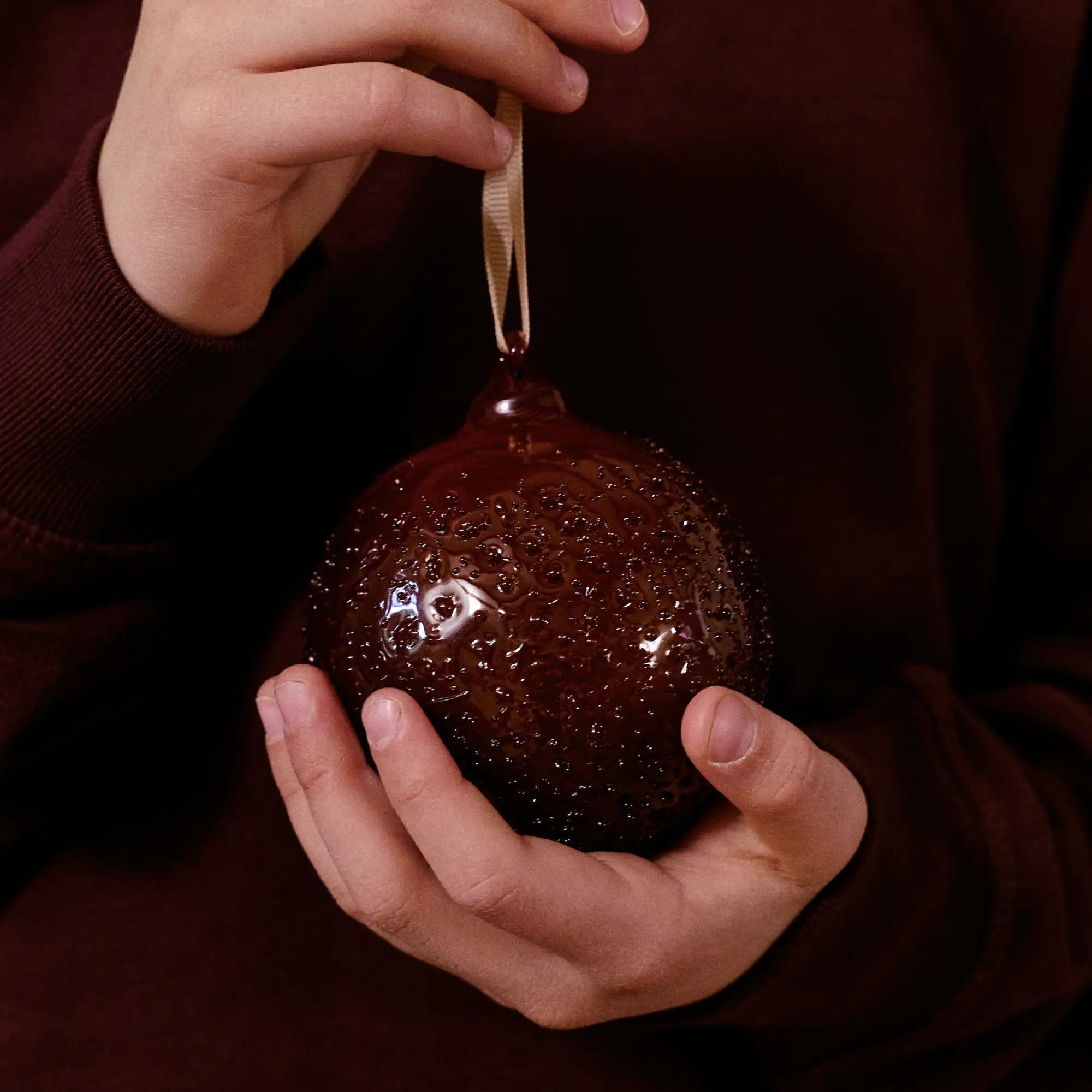 Julekollektionen, hænder der holder en mørkerød julekugle i boblet glas med båndophæng