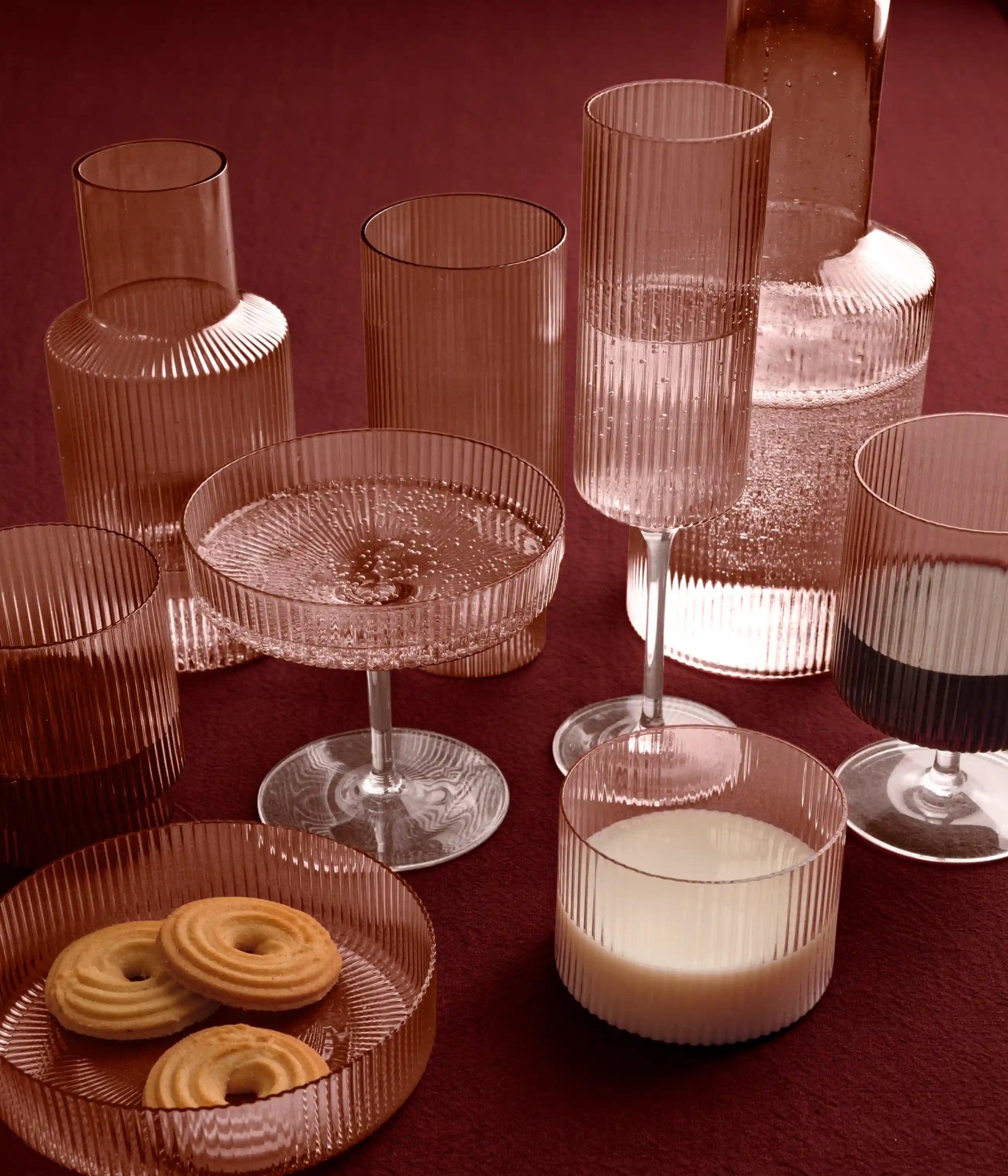 Riflet glas i rosa på et rødt bord, karaffel, glas, coupe og tallerken med kiks