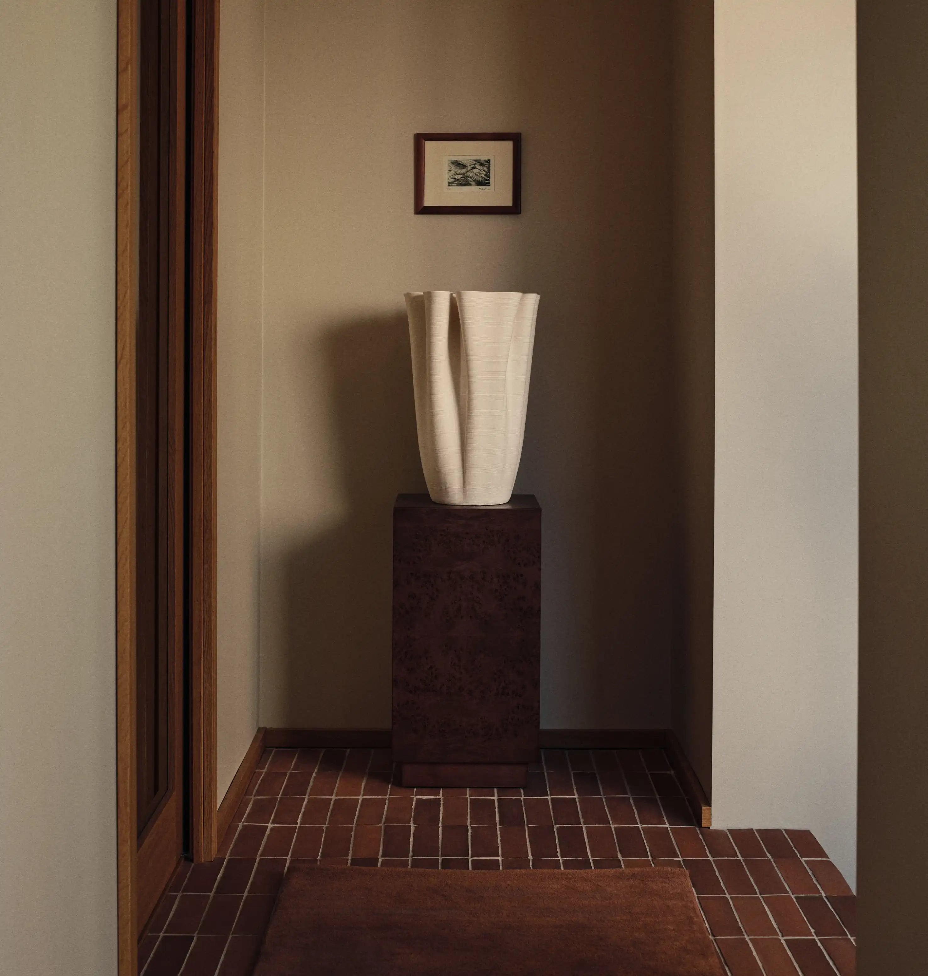 Entréen, høj off-white draperet vase på en mørk sokkel i en flisebelagt entré, lille indrammet tryk ovenover