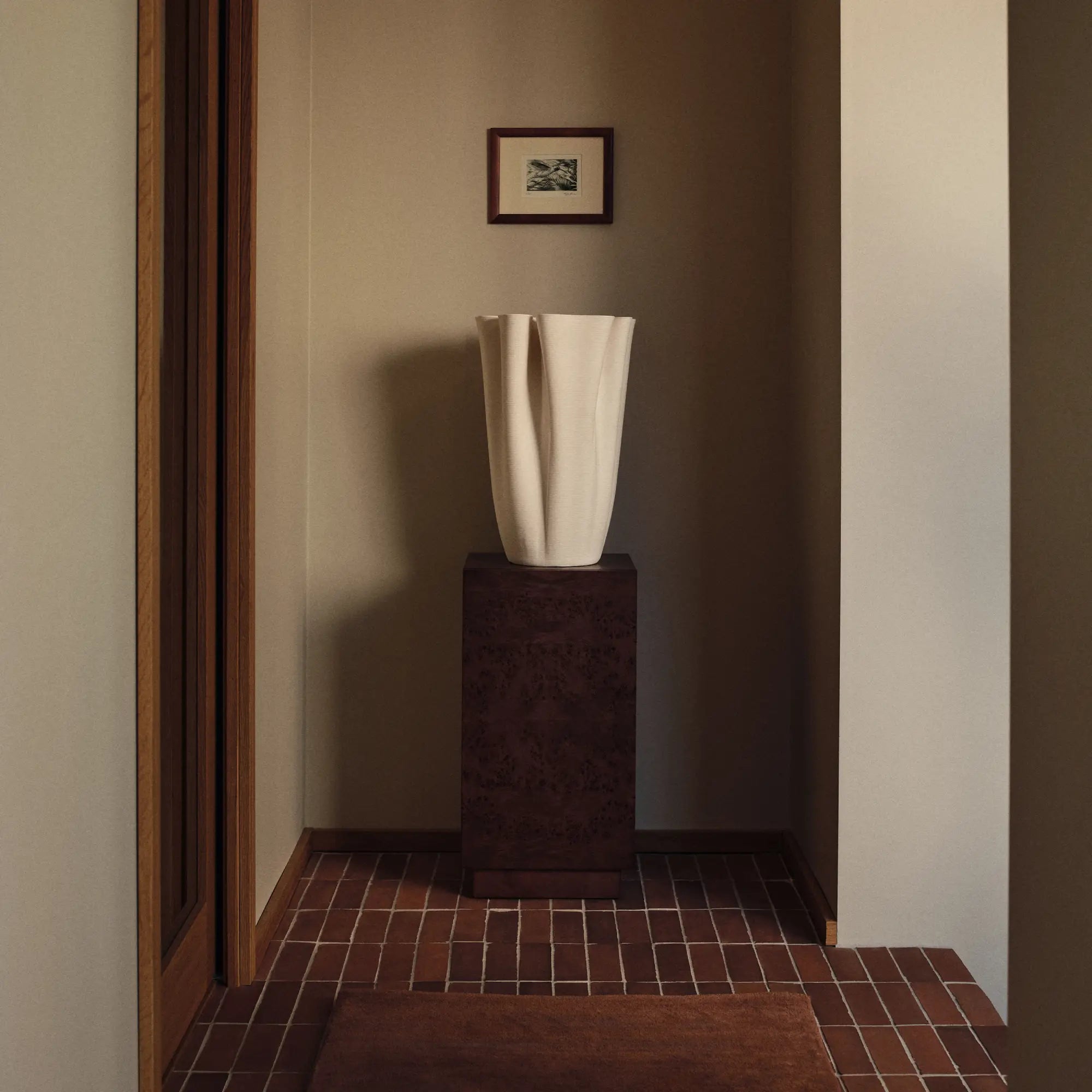 Entréen, høj off-white skulpturel vase på en mørk piedestal i en flisebelagt entré med indrammet tryk